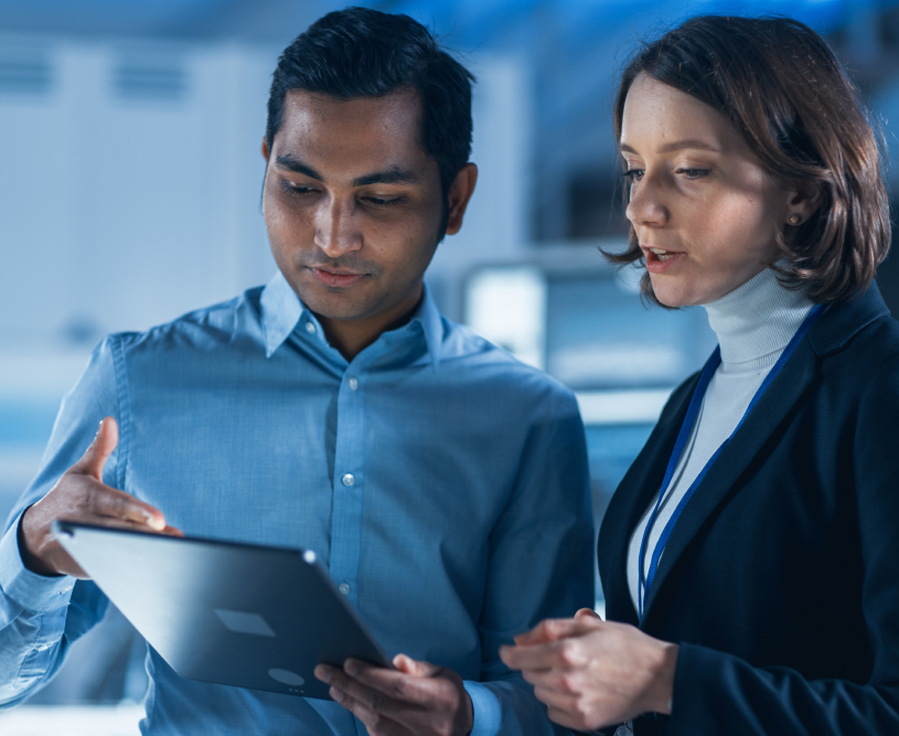 Two professionals reviewing information on a tablet in a modern office setting; a man in a light blue button-down shirt gestures toward the screen while a woman in business attire attentively listens.